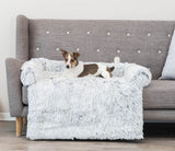Ein Hund liegt auf einer grauen Couch mit einem weißen, flauschigen Überwurf in einem Raum mit Holzboden.