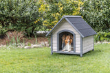 Ein Hund liegt in einer grauen Hundehütte auf einer grünen Wiese vor Büschen.