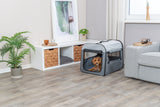 Ein Hund sitzt in einer grauen Transportbox in einem Wohnzimmer mit Holzfußboden, einem Sofa und Pflanzen.