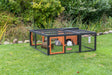 Kleintiergehege aus Metall mit Holzdetails auf grünem Rasen, innen zwei Kaninchen und Spielzeug.