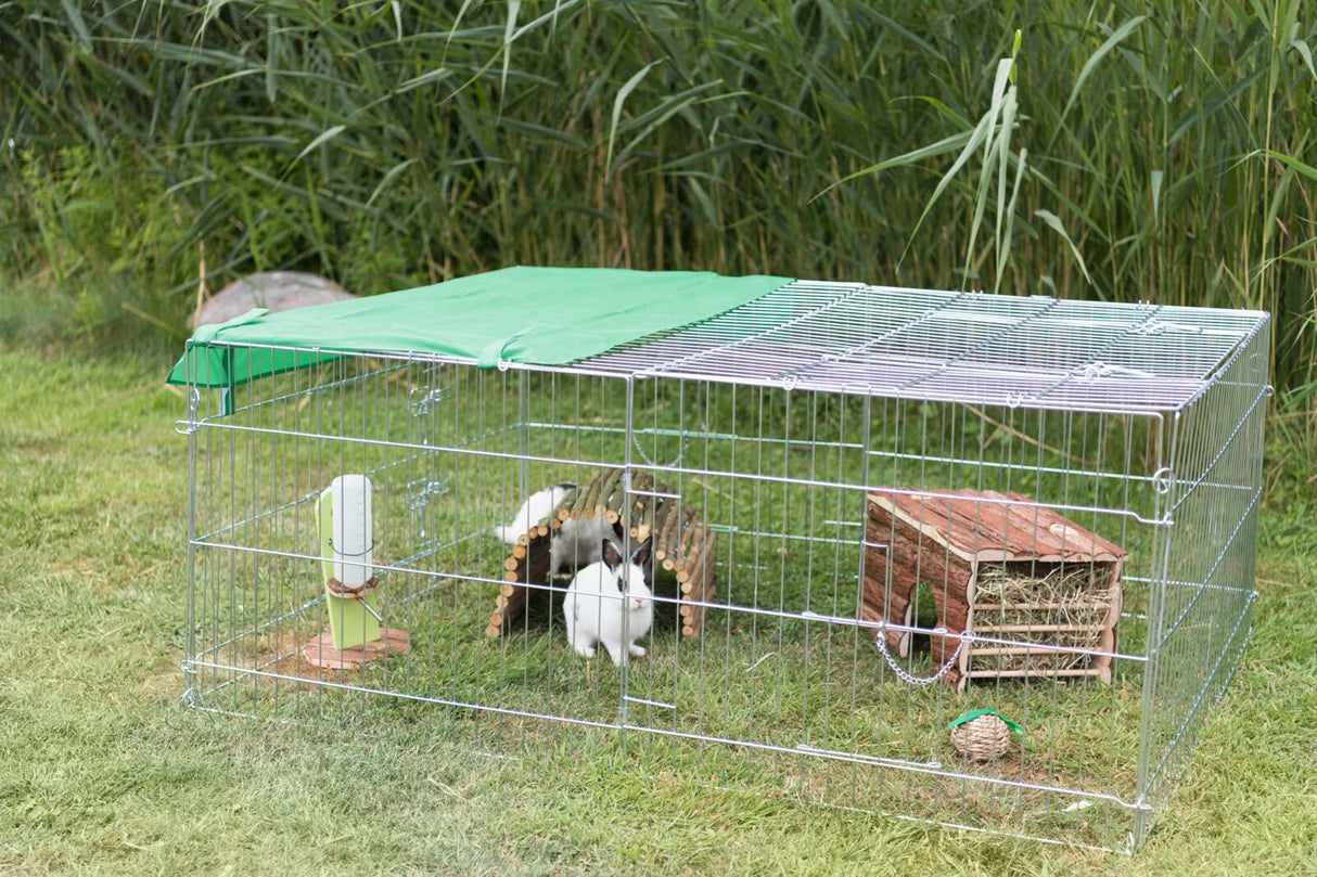 Ein rechteckiger Freilaufstall aus Metallgittern steht auf einer Wiese, darin sind Kaninchen und Spielzeug sichtbar.