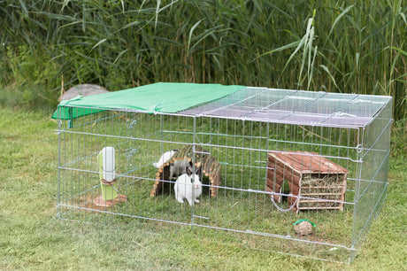 Ein rechteckiger Freilaufstall aus Metallgittern steht auf einer Wiese, darin sind Kaninchen und Spielzeug sichtbar.