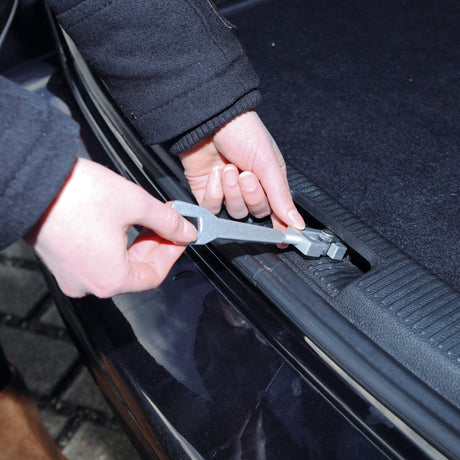 Eine Person hält ein metallisches Werkzeug und betätigt damit eine Schraube oder Mutter an einem schwarzen Kfz-Teil.