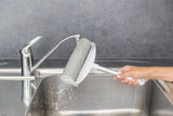 Hand hält Reinigungswerkzeug mit silbernem Griff unter laufendem Wasserhahn in Edelstahlspüle.