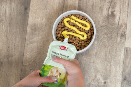 Hand hält TRIXIE Veggie-Topping über Hundenapf mit braunen Kroketten auf Holzboden.