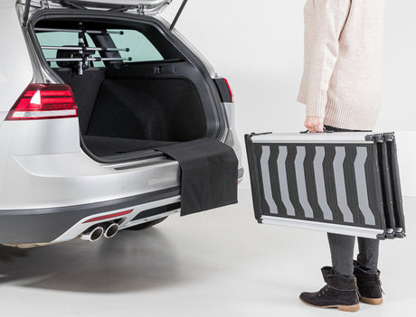 Eine Person steht neben einem geöffneten Kofferraum eines silbernen Autos und hält eine zusammenklappbare Rampe.