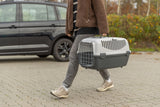 Person trägt graue Transportbox neben schwarzem Auto auf gepflastertem Weg.