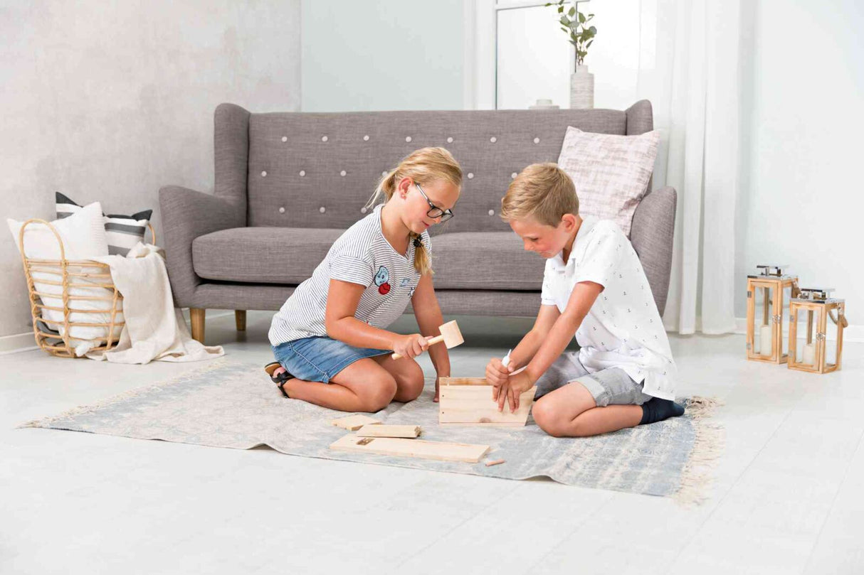 Zwei Kinder bauen gemeinsam eine Holzkiste auf einem Teppich vor einem grauen Sofa.