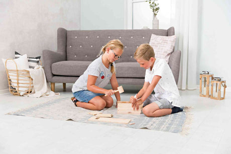 Zwei Kinder bauen gemeinsam eine Holzkiste auf einem Teppich vor einem grauen Sofa.