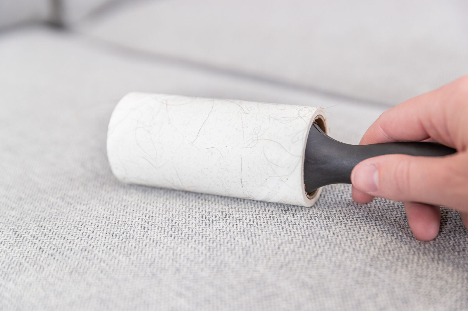Eine Hand hält eine schwarze Fusselrolle mit weißem Papier, die über eine graue Stoffoberfläche gerollt wird.