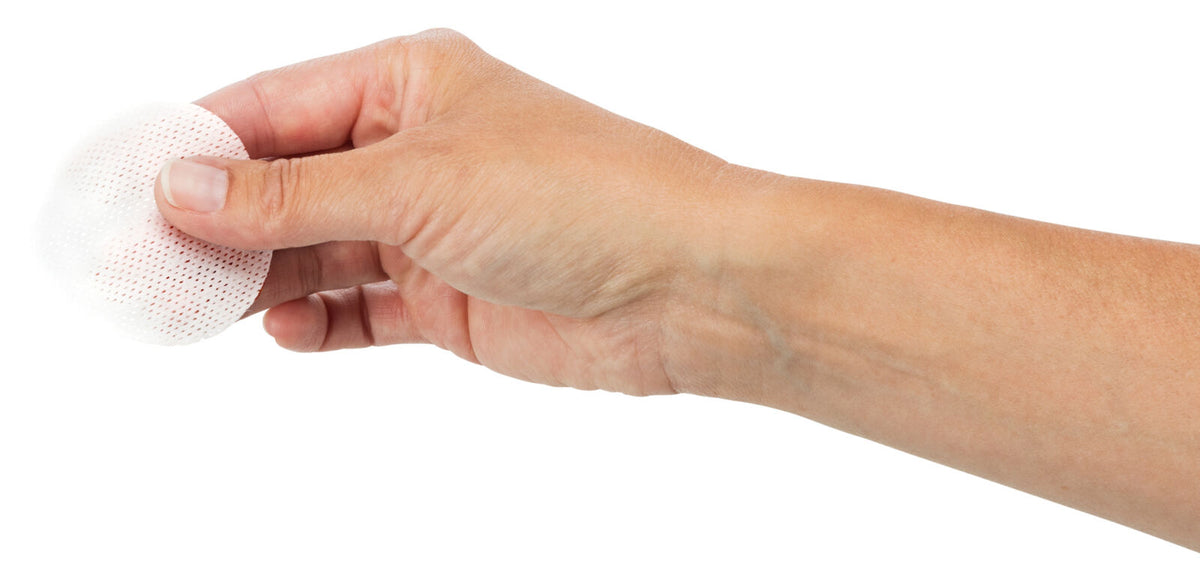Eine Hand hält ein rundes weißes, perforiertes Pad oder Tuch vor weißem Hintergrund, das zwischen Daumen und Zeigefinger fixiert ist.